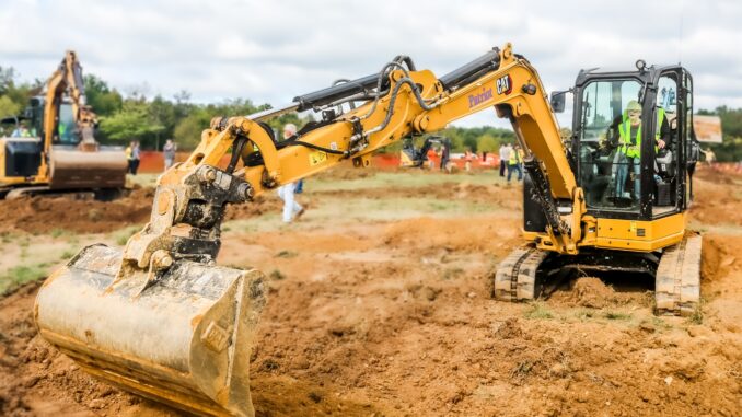 Yellow compact excavator working on construction site with other machinery in background.