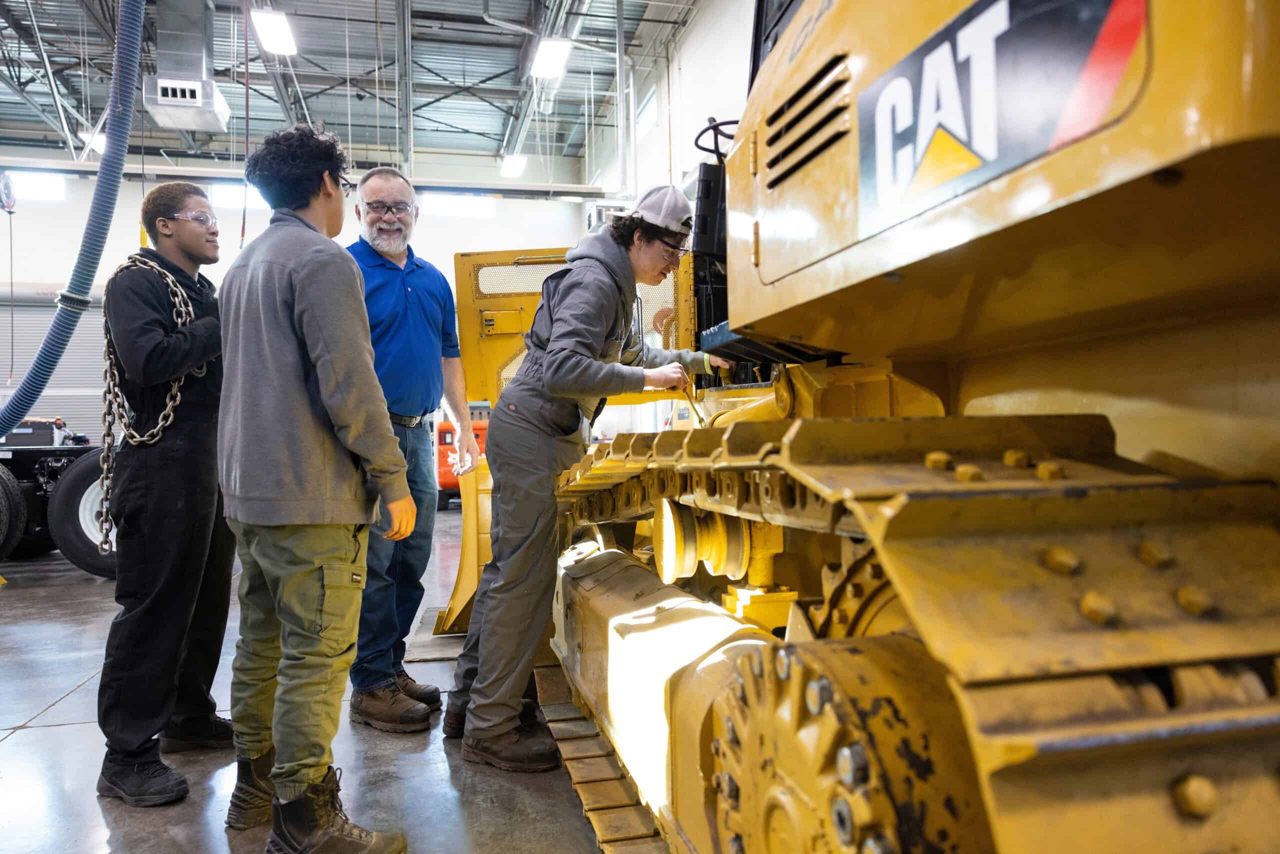 Group of students and instructor examining a yellow CAT construction vehicle in a training facility.