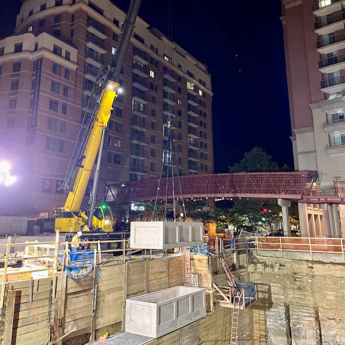 Nighttime construction site with crane lifting large concrete blocks between high-rise buildings.