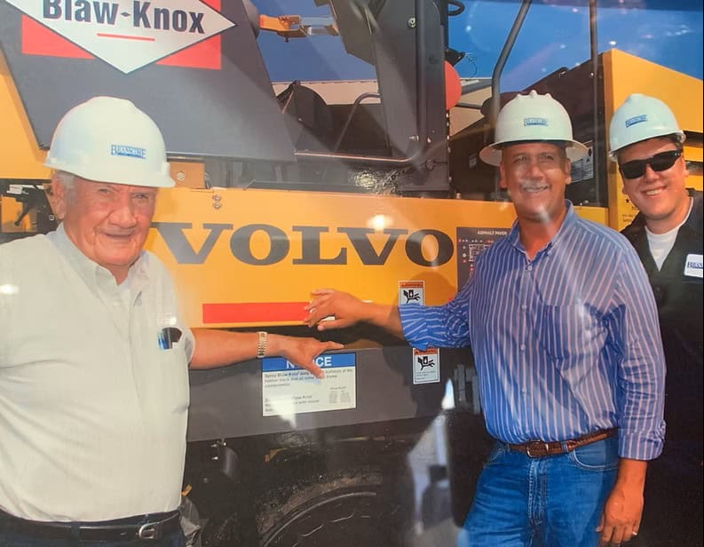 Three men in safety helmets standing in front of a Volvo construction vehicle.