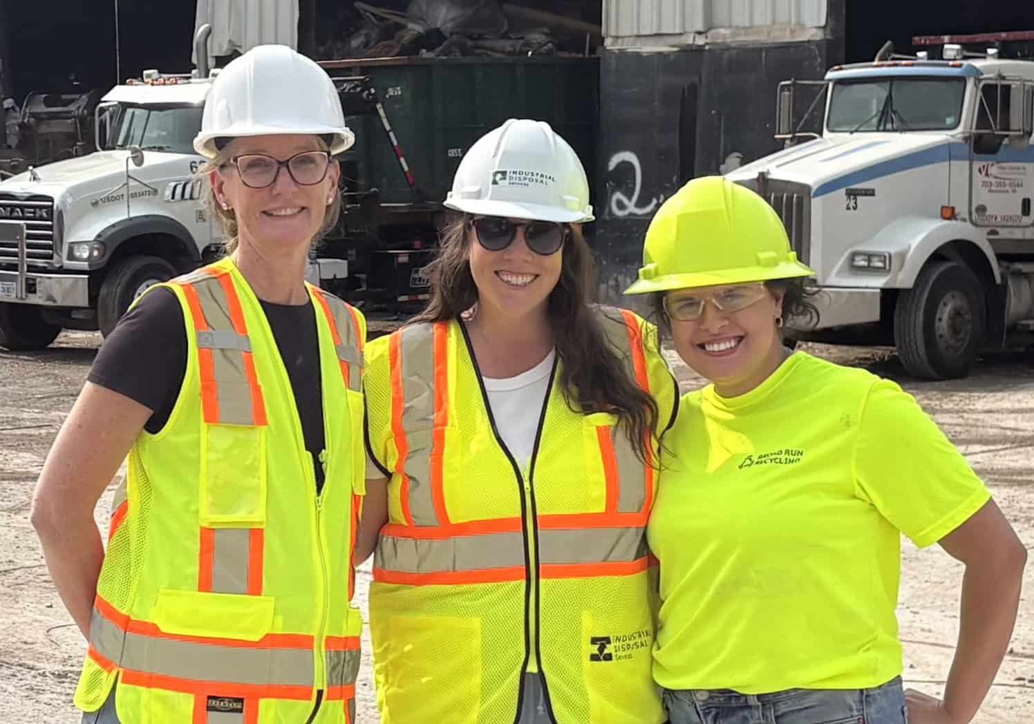 Three women in safety gear at a construction site with trucks in background.